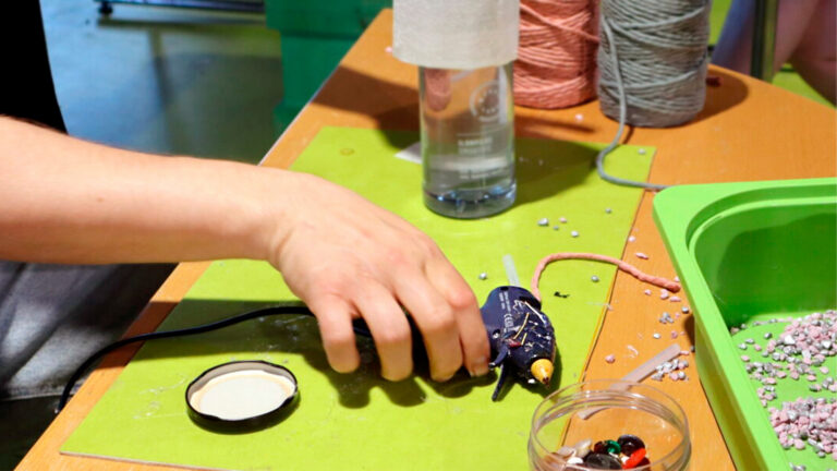 Korjaustorstain kävijä pitelemässä kuumaliimapistoolia, pöydällä näkyy korjausmateriaaleja kuten lankaa ja helmiä