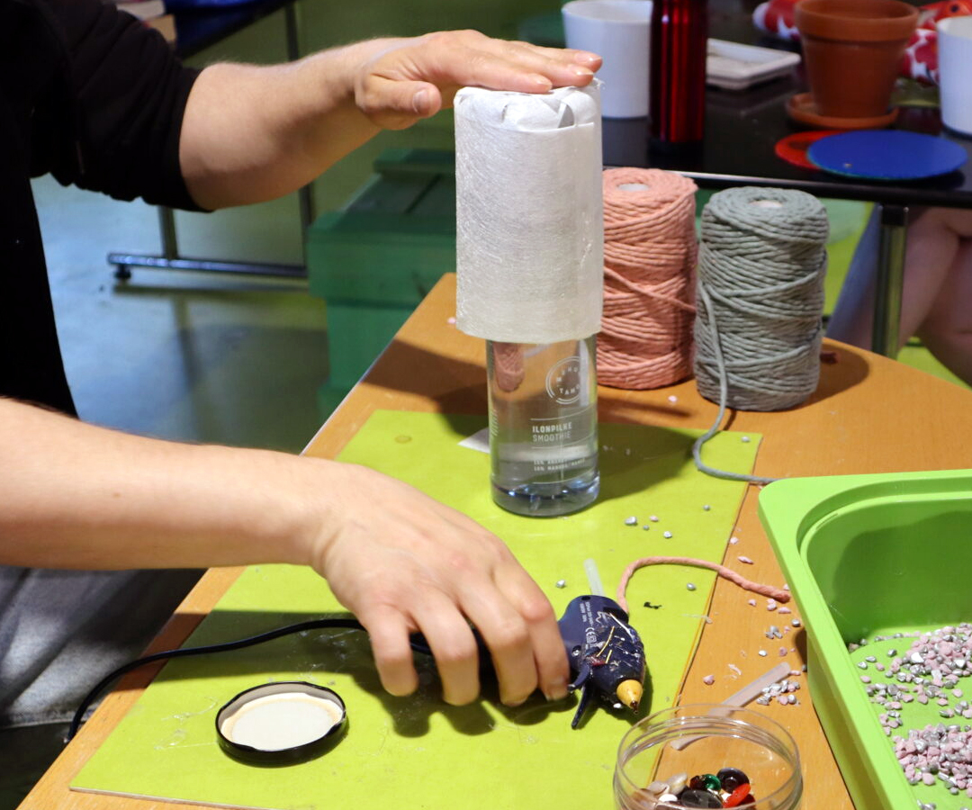 Korjaustorstaina pöydälle levitettyjä korjausmateriaaleja sekä kuumaliimapistooli, ihmisen kädet pitelevät niitä.