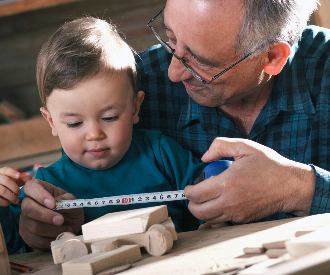 Isoisä pitämässä lapsen edessä avointa mittanauhaa, jolla opettaa lasta mittaamaan puista rakennuspalaa
