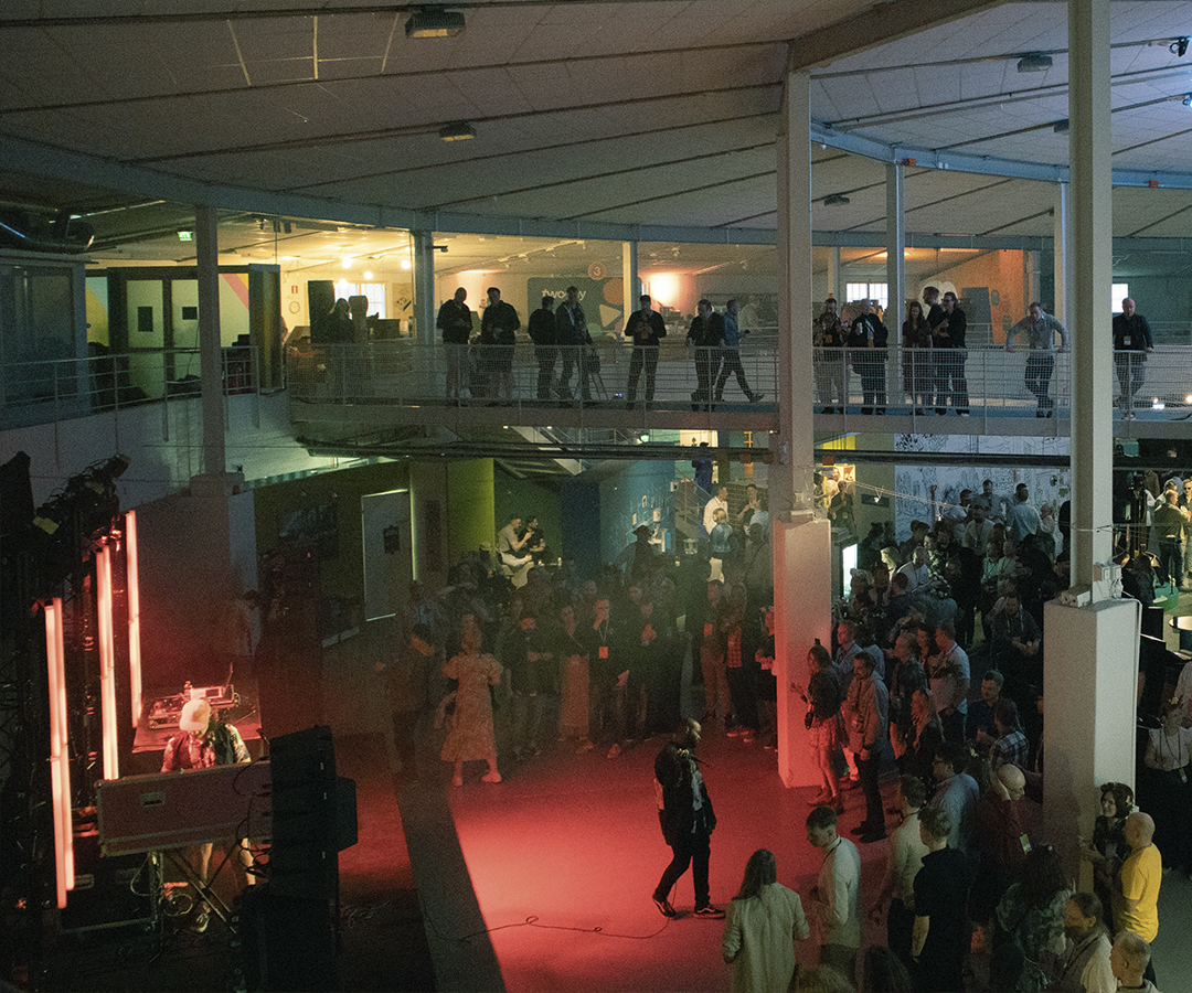Kuva Tekniikan museon päänäyttelyhallista, jossa on käynnissä musiikkiesitys, lavan edessä mikrofonia pitelevä esiintyjä ja juhlivia ihmisiä