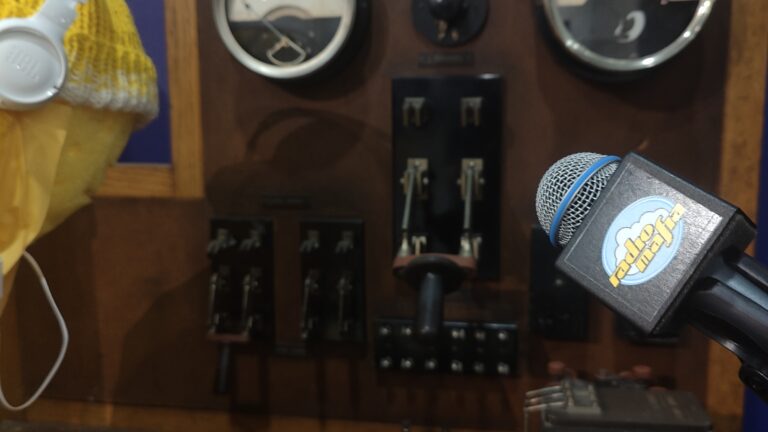 Keltainen ihmishahmo kuulokkeet päässä häärii kipinälennättimen äärellä. Vieressä seisoo Yleisradion Radio Mafian mikrofoni.